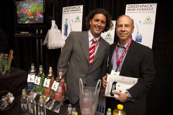 Two men in jackets pose at the Greenpack booth, in front of Greenpack New Format banners and a table of bottled drinks with tags.