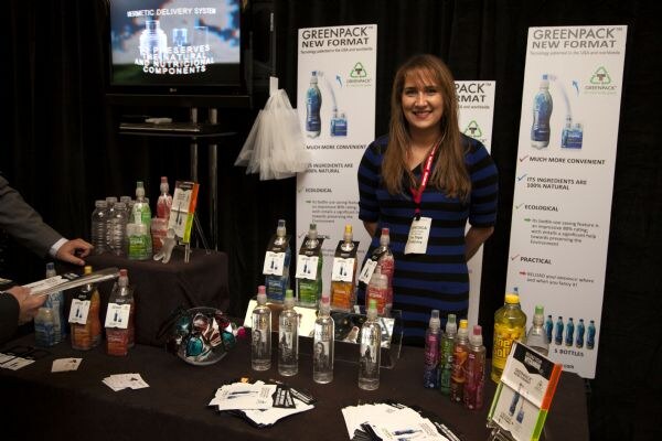 A smiling woman with long hair and a lanyard stands behind a table of bottled drinks and flyers, in front of Greenpack New Format banners and a monitor.