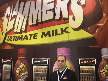 A man in a black shirt with a lanyard stands in front of a Slammers Ultimate Milk sign, with Slammers bottle graphics and lit refrigerated coolers behind him.