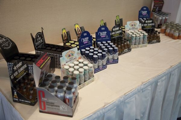 A long table with a white cloth holds rows of small shot bottles in display boxes, including a G-Tox box in the foreground and various sleep and recovery shots behind it.