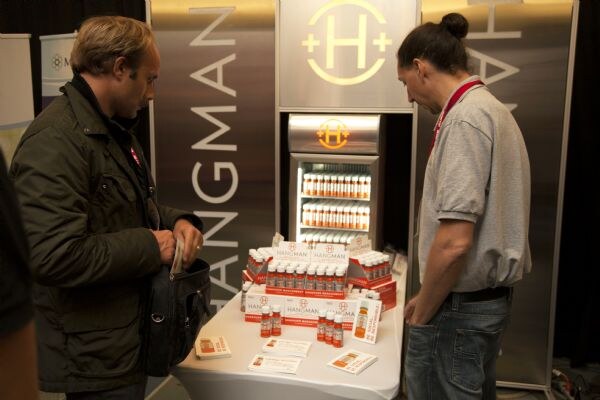 Two men stand at a Hangman booth with a table of small bottles and a lit refrigerator; one man looks into a bag while the other, wearing a red lanyard, looks at the display.
