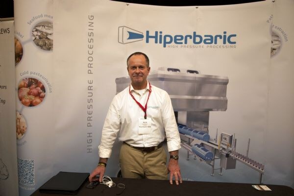 A man in a white shirt and red lanyard stands behind a table in front of a Hiperbaric high pressure processing backdrop showing processing equipment.