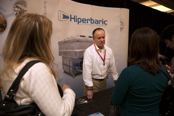 A man in a white shirt and red lanyard talks with two women seen from behind in front of a Hiperbaric backdrop showing a high pressure processing machine.