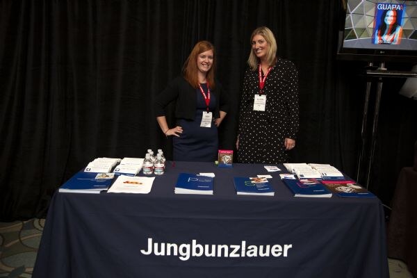 Two women with red lanyards stand behind a navy table marked Jungbunzlauer, which holds brochures, folders and small water bottles in front of a black curtain.