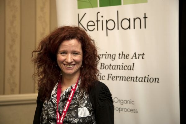 A smiling woman with curly red hair and a red lanyard stands in front of a Kefiplant banner.
