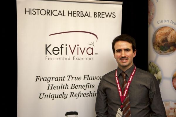 A smiling man with a mustache, wearing a tie and red lanyard, stands beside a KefiViva banner reading Historical Herbal Brews, Fermented Essences.