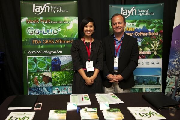 A woman and a man in dark jackets with red lanyards stand behind a table of brochures in front of layn Natural Ingredients banners, one promoting Go-Luo monk fruit.