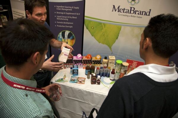 A man gestures toward a table of bottles and cartons beneath a MetaBrand banner while two attendees, seen from behind, listen.