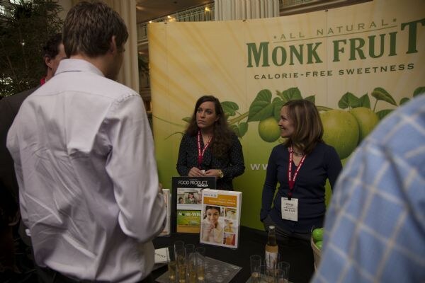 Two women with lanyards stand behind a table in front of a green All Natural Monk Fruit banner, while two men in the foreground face them. Magazines and small glasses sit on the table.