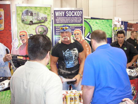 A man with a blond mustache and sunglasses on his head stands in a Socko booth, with a Why Socko sign and Socko graphics behind. Other attendees stand around him.