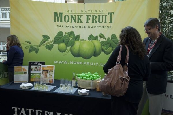 Monk Fruit display with a green banner reading All Natural Monk Fruit, with a bowl of green fruit and small cups on a black table. A woman and a man with a lanyard stand nearby.