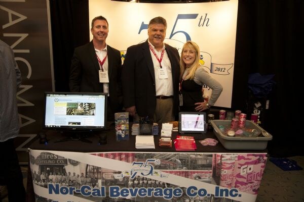 Two men and a woman in business attire stand behind a Nor-Cal Beverage Co. Inc. table with a monitor, a tablet, and pink cans, in front of a 75th anniversary sign.