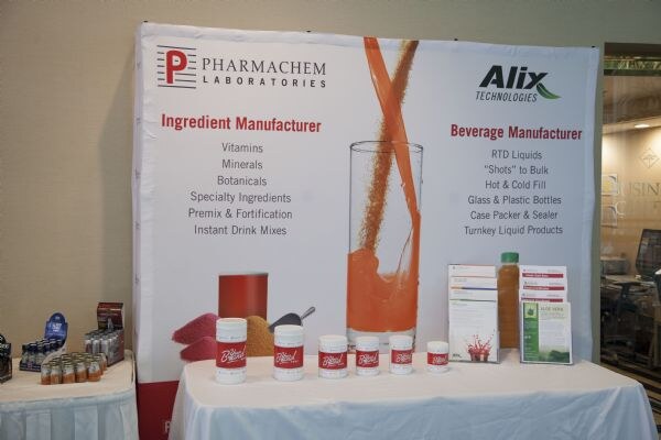 A Pharmachem Laboratories and Alix Technologies backdrop listing ingredient and beverage manufacturing, with white product tubs and sample bottles on a draped table.