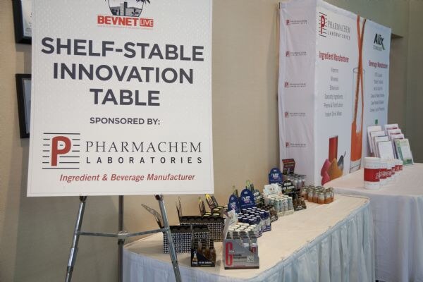 A sign reads Shelf-stable Innovation Table, sponsored by Pharmachem Laboratories. Behind it, a table with a white skirt holds assorted small bottles and shot packaging.