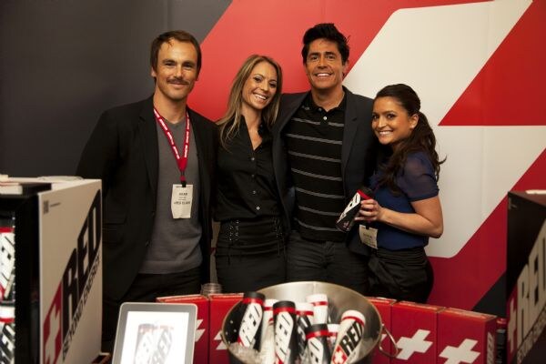 Two men and two women stand close together smiling behind a table with a bucket of Red cans and red boxes with white crosses. A Red sign is at left and a red and white backdrop is behind.