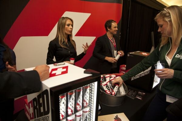 A woman and a man in dark clothes talk at a Red booth beside a lit Red cooler and a bucket of cans. A woman in a green jacket at right reaches toward the cans holding a clear cup.