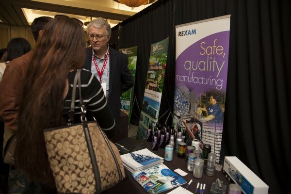 Attendees talk at the Rexam booth. A gray-haired man with glasses and a lanyard stands behind a table of brochures and cans, beside a Rexam banner reading Safe, quality manufacturing.