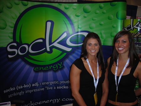 Two smiling women with long dark hair, wearing black tops and lanyards, stand in front of a green and blue Socko Energy banner.