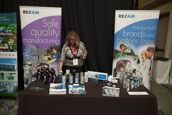 A woman with a lanyard stands behind a dark table of brochures, cans and bottles, between two Rexam banners reading Safe, quality manufacturing and Helping build brands and bottom lines.