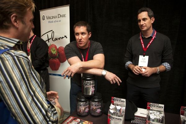 Three men talk at a table with tubs and packets of Xtend product, beside a Virginia Dare banner showing strawberries. One man in a striped shirt faces away from the camera.