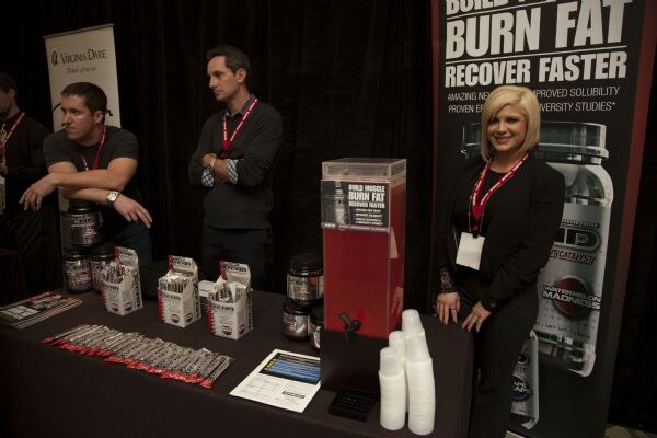 Two men and a blonde woman wearing red lanyards stand at a table of supplement tubs and sample packets, in front of a Virginia Dare banner and a Burn Fat Recover Faster banner.