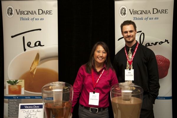 A smiling woman in a magenta shirt and a man in a black shirt stand between two Virginia Dare banners, behind two beverage dispensers of tea.