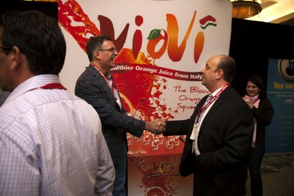 Two men in dark jackets and red lanyards shake hands and smile in front of a ViVi orange juice banner. A man in the foreground and a woman in the background look on.