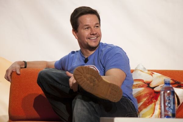 A smiling man in a blue t-shirt and jeans sits on an orange couch with one foot raised and a lapel microphone. A bottle of AQUA water sits on the table in front of him.