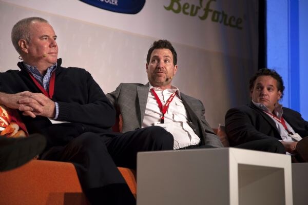 Three men in jackets and red lanyards sit on stage chairs, two wearing headset microphones, under a BevForce banner.