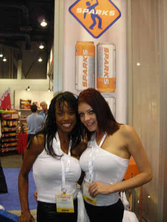 Two smiling women in white tops and badge lanyards stand in front of a Sparks banner showing two Sparks cans, with a snack display and other attendees behind them.