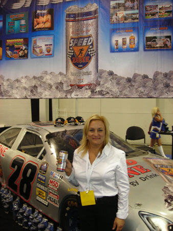 A blonde woman in a white shirt holds a can of Speed Energy Drink beside a race car numbered 28 with MSD decals, under a large Speed banner.