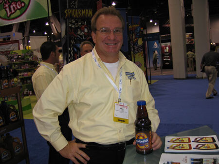 A smiling man with glasses, a yellow shirt and a badge stands beside a table with a brown bottled drink. A Spiderman sign hangs behind him in the convention hall.
