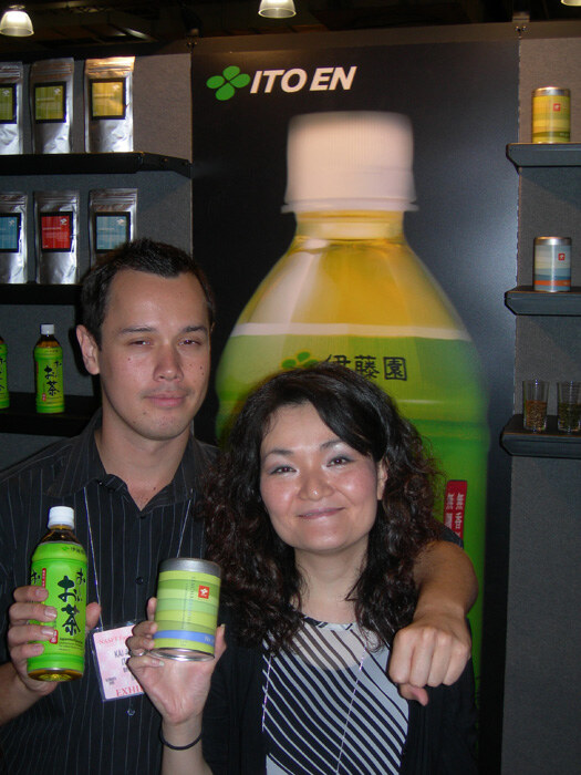 A man and a woman pose close together in front of an Ito En sign with a large green tea bottle graphic. He holds a bottled green tea and she holds a tea tin; tea packages sit on shelves behind.