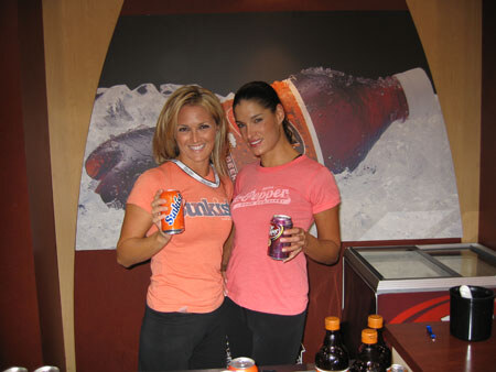 Two smiling women in orange and pink shirts stand behind a counter. One holds a Sunkist can and the other a Dr Pepper can, in front of a large bottle graphic.