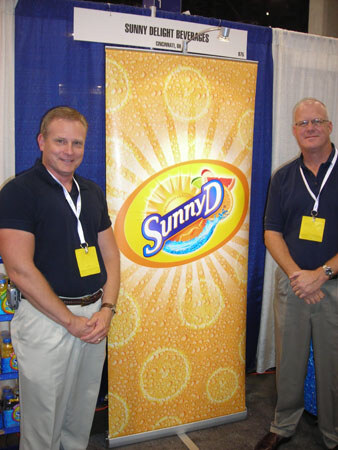 Two men in dark polo shirts with yellow badges stand on either side of a tall SunnyD banner under a Sunny Delight Beverages, Cincinnati, OH sign.