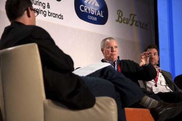 Seated panelists on stage, one gesturing, talk with a man in the foreground facing away, beneath banners for Crystal Cooler and BevForce.