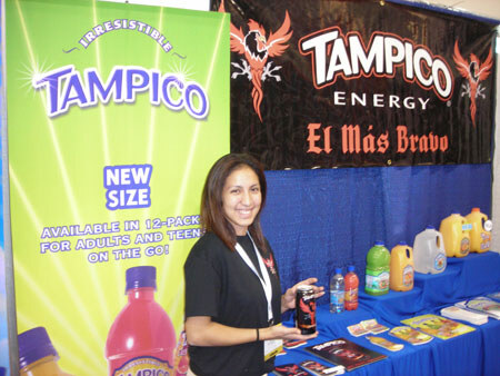 A smiling woman with long dark hair holds an energy drink can at a booth with a green Tampico banner, a black Tampico Energy 'El Más Bravo' banner, and juice bottles on a blue-draped table.