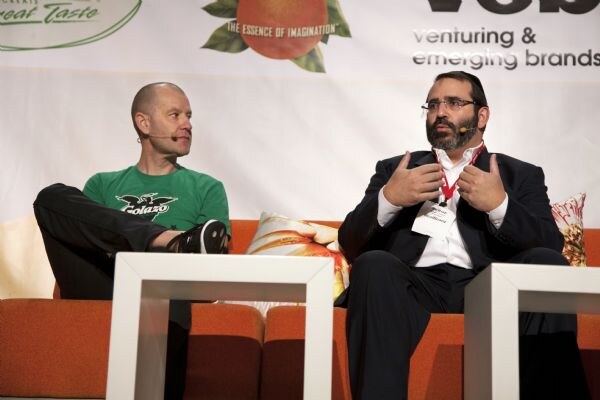 Two men sit on an orange couch on stage. One wears a green Golazo shirt and a headset microphone; the other, in a dark suit and glasses, gestures with both hands as he speaks.