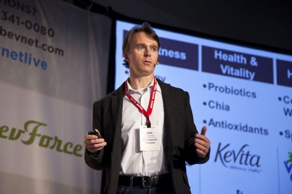 Mike, wearing a badge and a red lanyard, speaks on stage with a headset microphone in front of a projected slide listing probiotics, chia and antioxidants, with a KeVita logo.