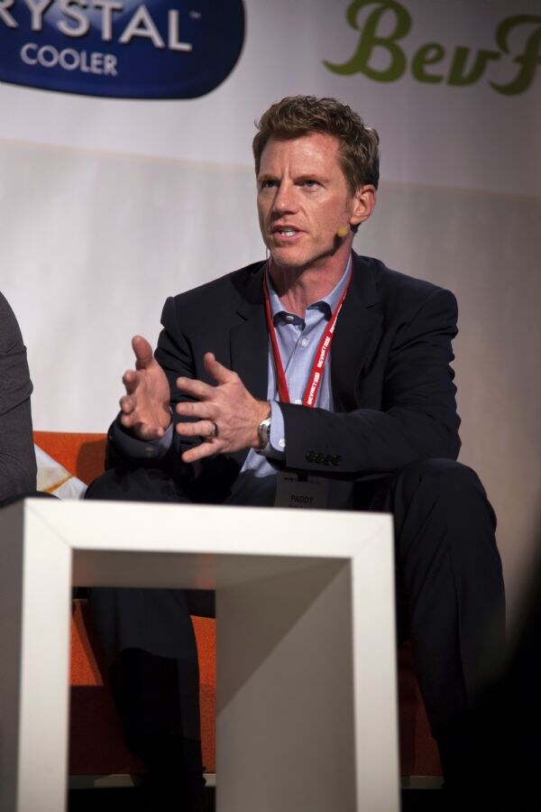 A man in a dark blazer and light blue shirt with a headset microphone and a Paddy badge gestures while seated on stage in front of a Crystal Cooler banner.