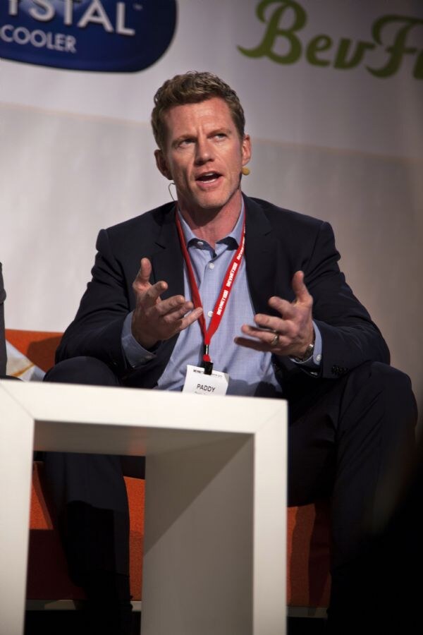 A man in a dark blazer and light blue shirt with a headset microphone and badge reading Paddy gestures with both hands while seated on stage.