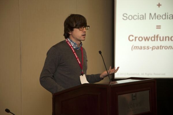 A man with glasses and a gray sweater speaks at a podium, gesturing toward a slide reading Social Media = Crowdfund, with a RocketHub copyright line.