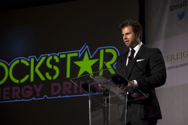 A man in a dark suit and tie speaks at a clear acrylic podium on a stage, beside a large screen showing the Rockstar Energy Drink logo.