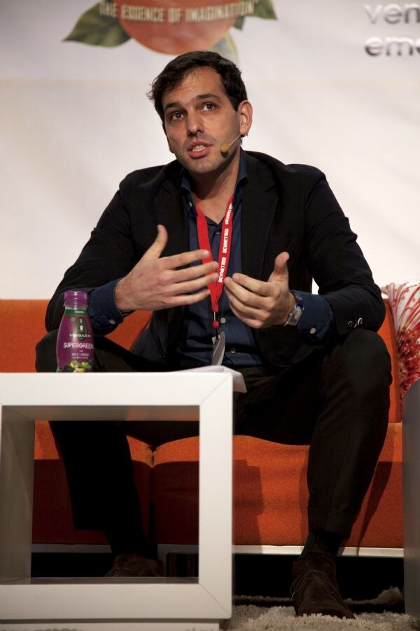 A man in a dark jacket with a red lanyard and headset microphone gestures while seated on an orange couch. A purple Sambazon Supergreen bottle sits on the white table in front of him.