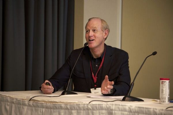 A man with gray hair in a dark blazer and red lanyard, badge reading Scott, gestures while speaking at a draped table with two microphones and a water bottle.