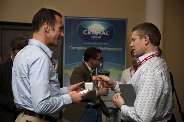 Two men in dress shirts and lanyards talk and exchange small items in front of a Crystal Cooler banner, with other attendees behind them.