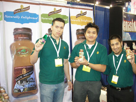 Three men in green Taste Nirvana polo shirts and badges stand in front of banners and a tall bottle graphic of Taste Nirvana Thai Mulberry Jasmine Tea; two hold small bottles.