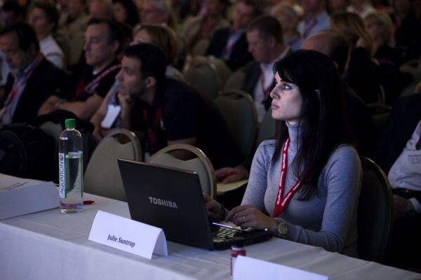 A woman with long dark hair and a red lanyard looks to the side while seated at a table with a Toshiba laptop, a water bottle and a name card. Other attendees sit behind her.