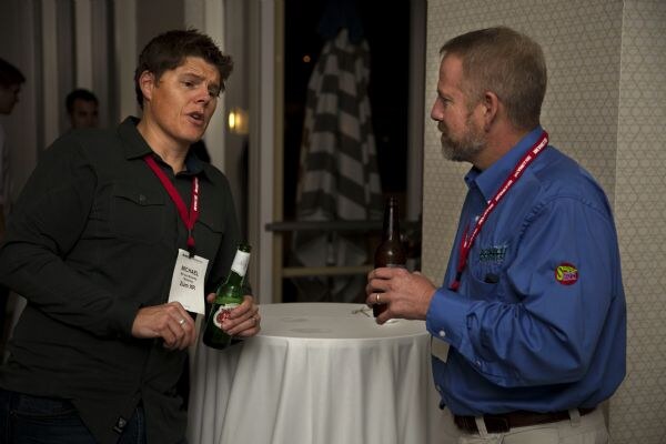 Michael, on the left, wears a red lanyard and holds a green beer bottle while talking with a bearded man in a blue shirt holding a bottle, beside a white-draped cocktail table.