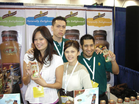 Four people smile in front of Taste Nirvana banners reading Naturally Enlightened. Two women hold small bottles, and bottles are displayed along the bottom.